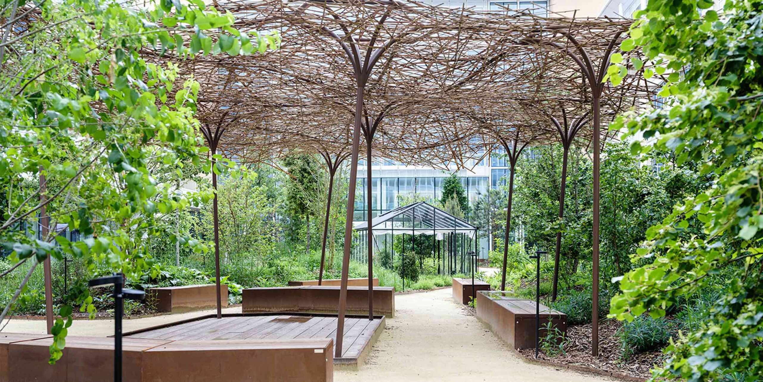 Anlagt grønt gårdrum med flettet pergola, cortenstål-bede og siddeplatform mellem bygninger.