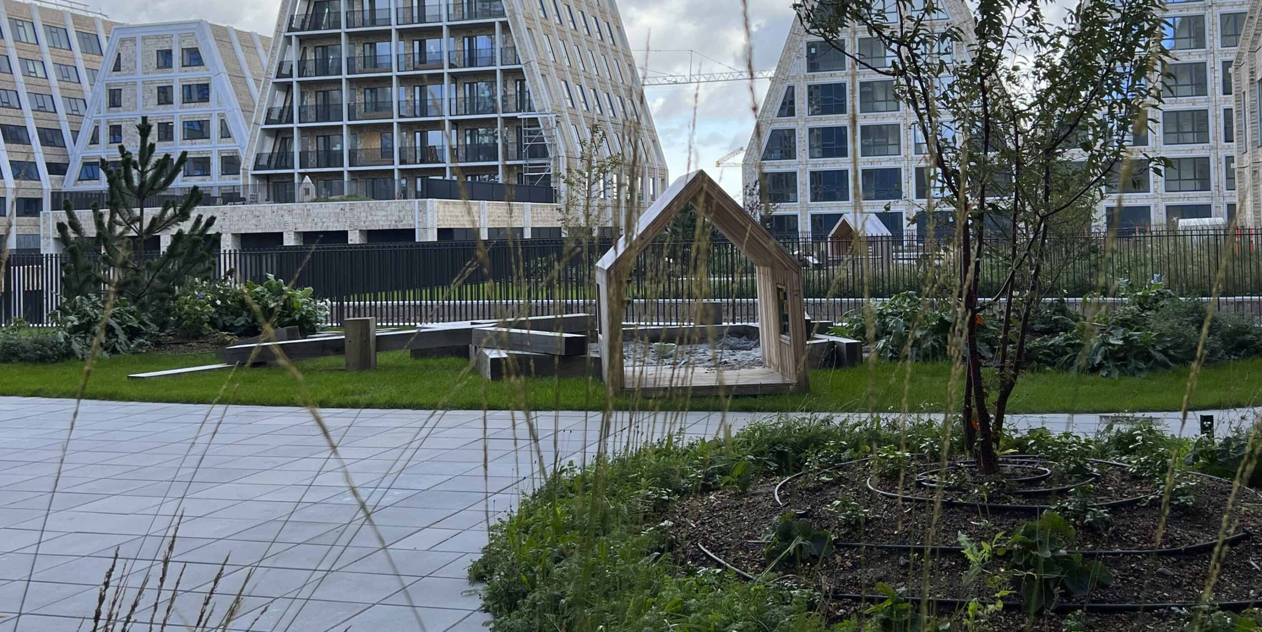 Nyanlagt gårdmiljø med legehus i træ, beplantning og græsplæne foran boligbebyggelse med karakteristiske trekantede facader.