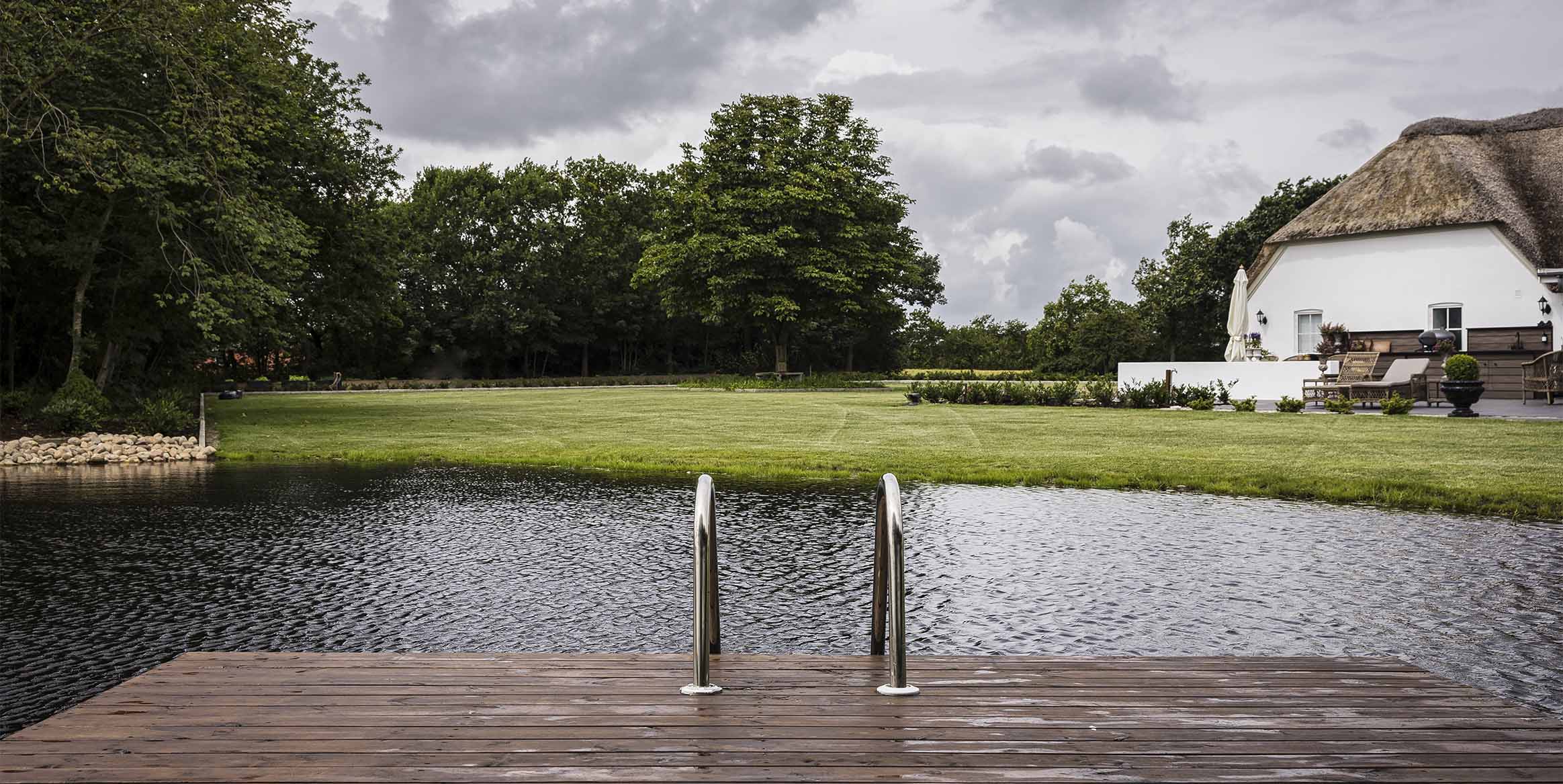 Badebro med stigetrin ved anlagt sø omgivet af plæne og stråtækt hus