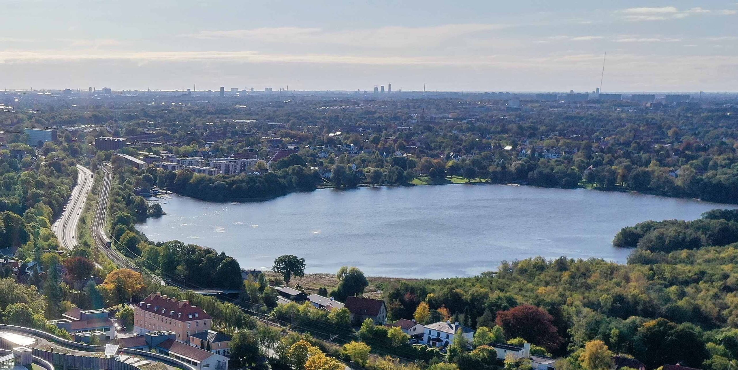 Luftfoto af sø omgivet af grønne områder og boligbebyggelse med bymidte i baggrunden.