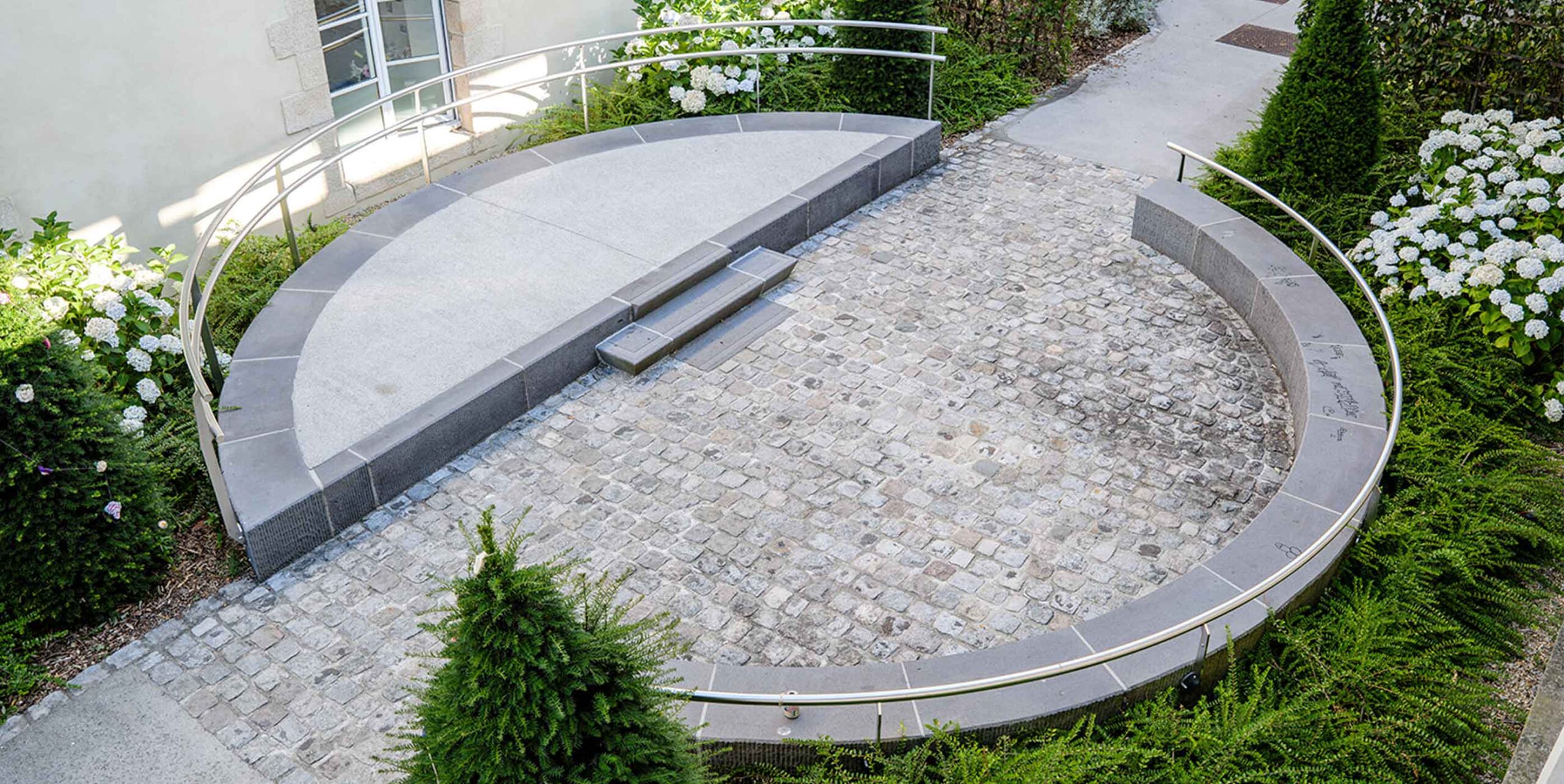 Cirkulær granitbelagt terrasse med trapper og rustfrit stålgelænder set fra oven, omgivet af hortensia og stedsegrønne buske.