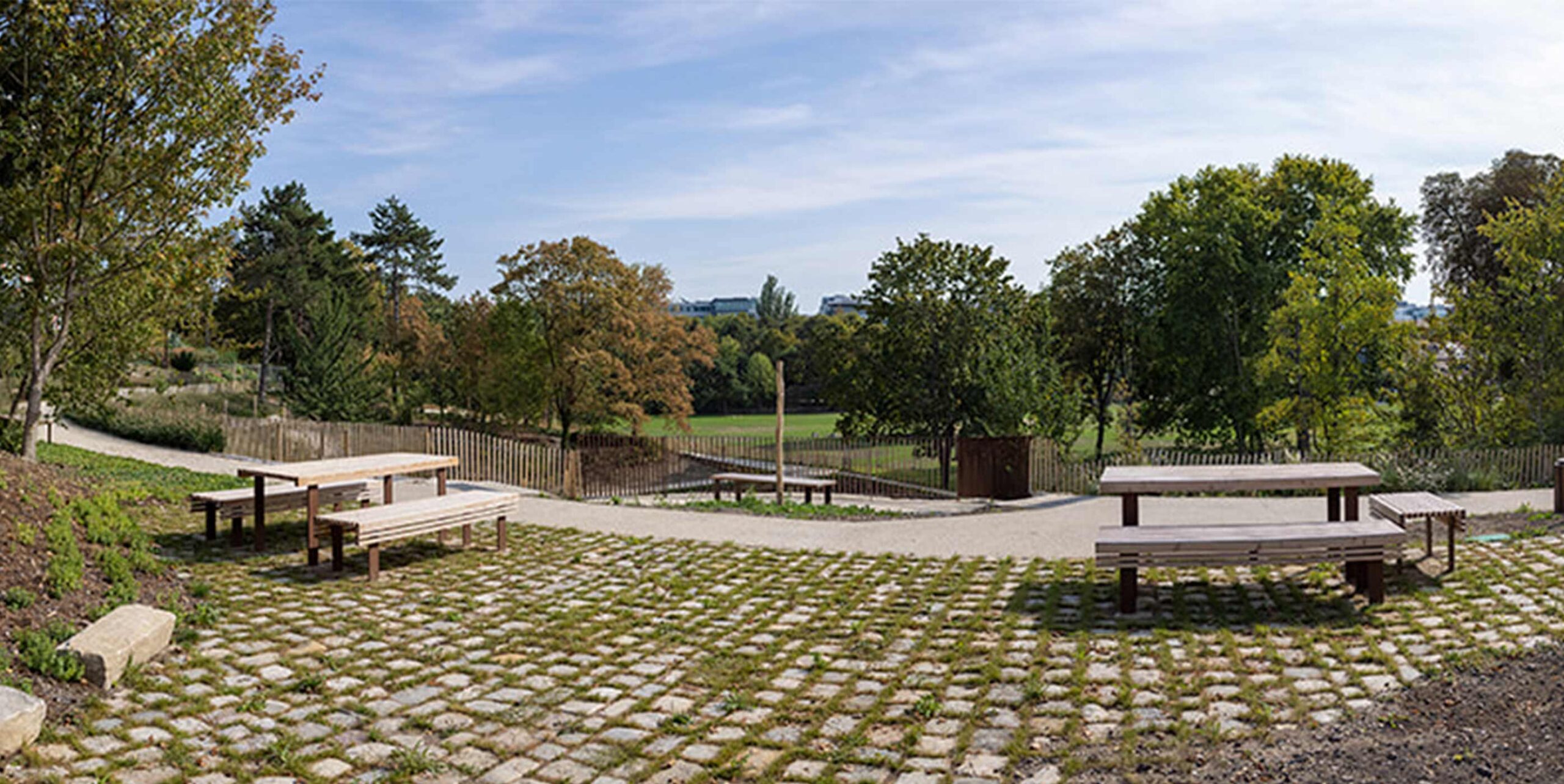 Anlagt opholdsområde i park med picnicborde på græsarmeret belægning omgivet af træer.