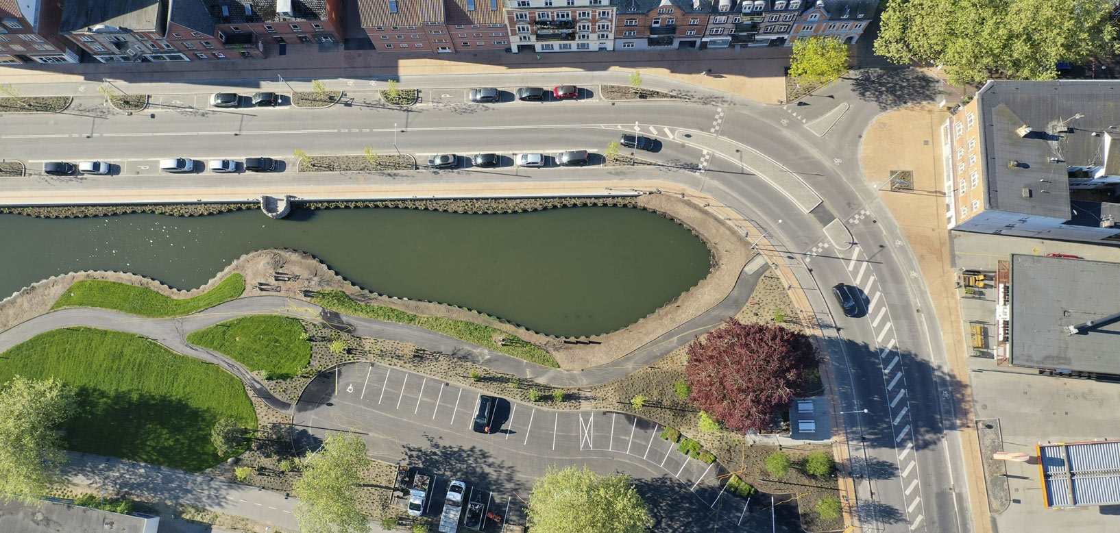 Luftfoto af nyanlagt bassin med grønne skråninger, stiforløb og parkeringsplads i bymiljø.