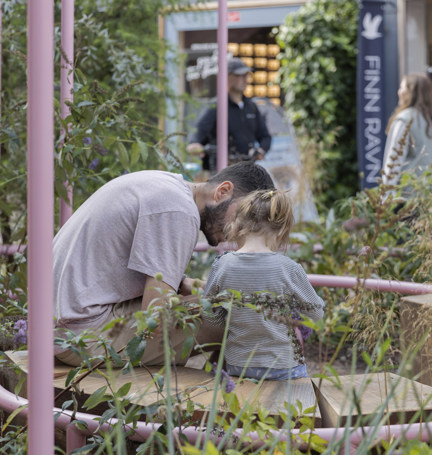 Voksen og barn undersøger planter sammen i bynært haveanlæg med træterrasse og beplantning.