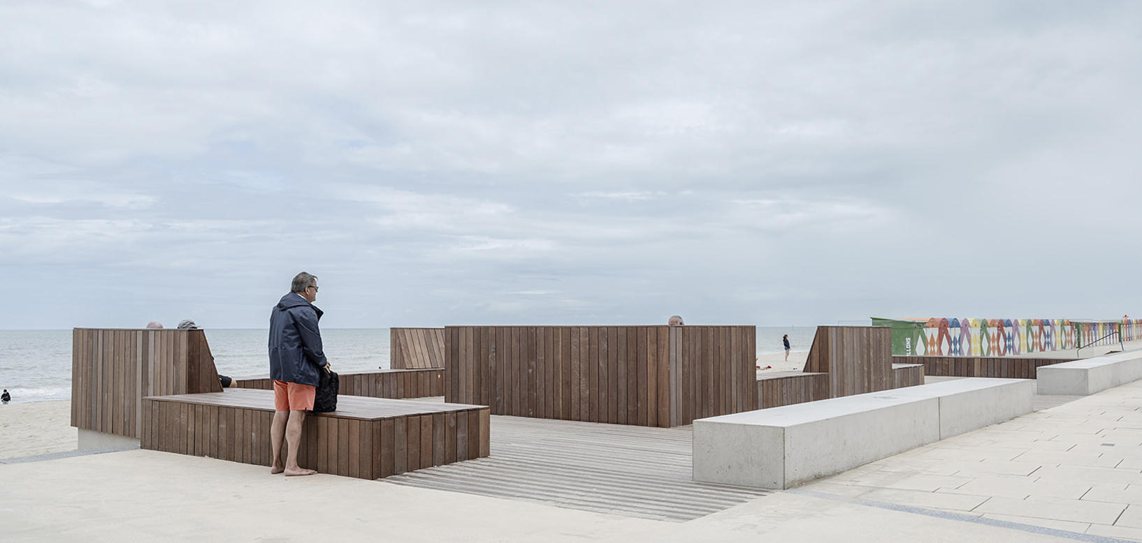 Træbeklædte læskærme og siddeområder på strandpromenade med udsigt til havet.