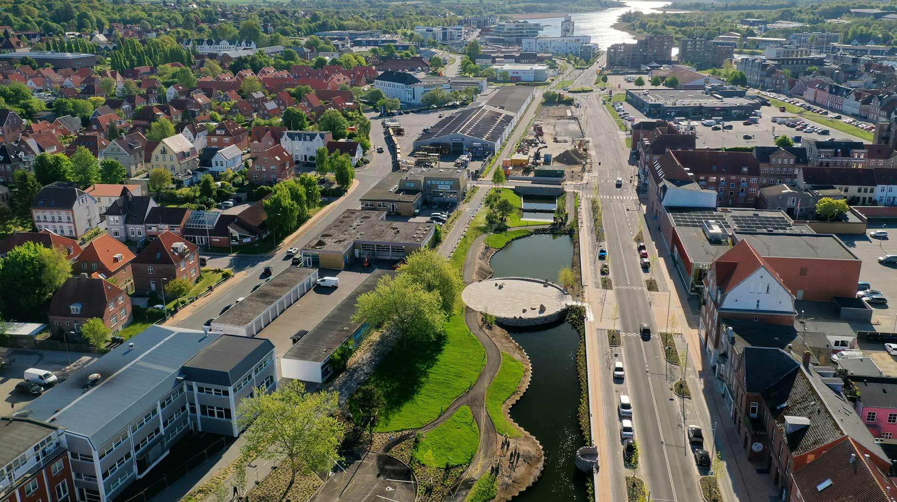 Luftfoto af nyanlagt klimatilpasningsprojekt med vandløb, grønt areal og stiforløb i by nær havn.