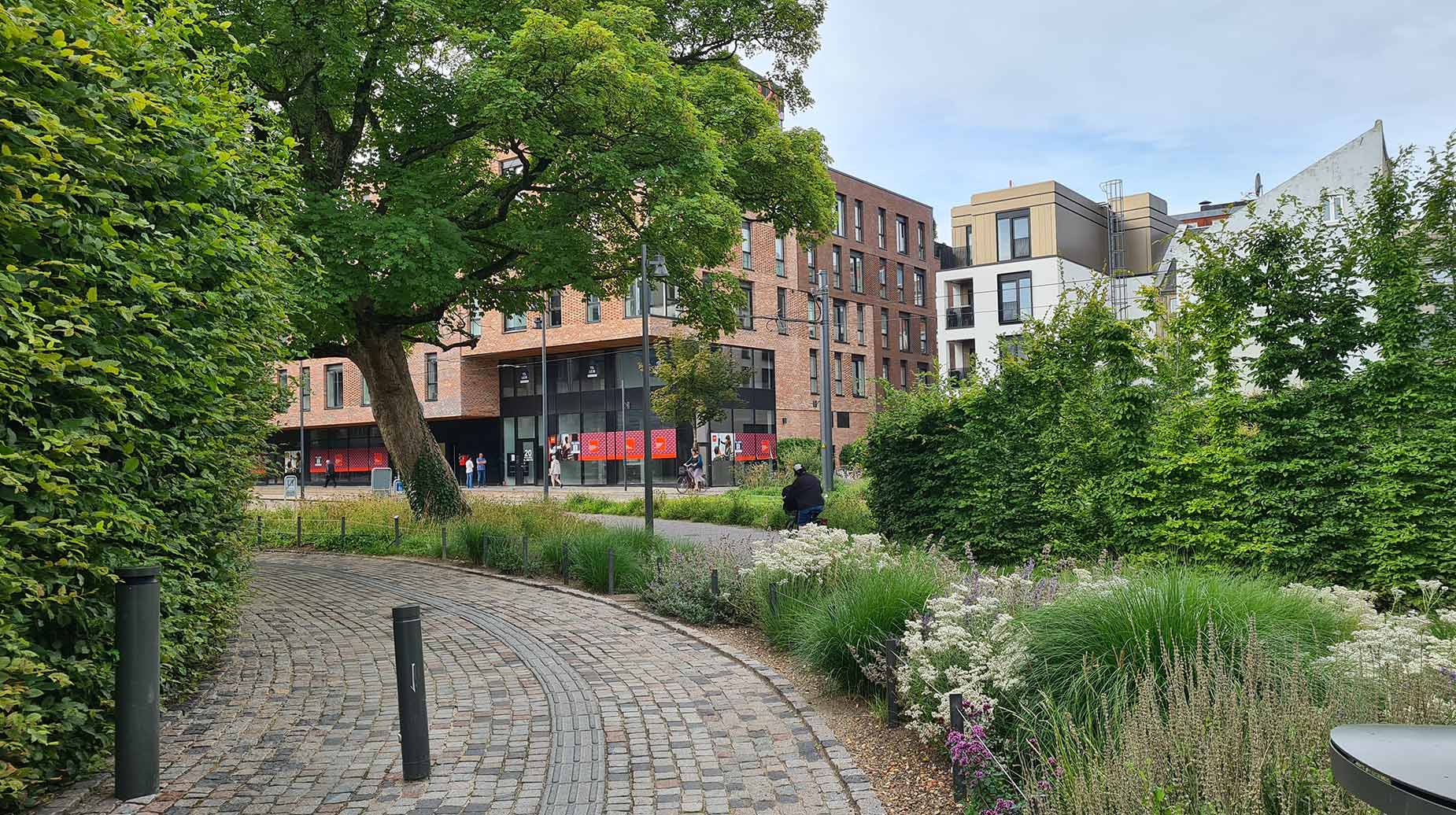 Brolagt gangsti gennem grønt byrum med stedsegrønne hække og blomstrende staudebede foran nybyggede boligejendomme.