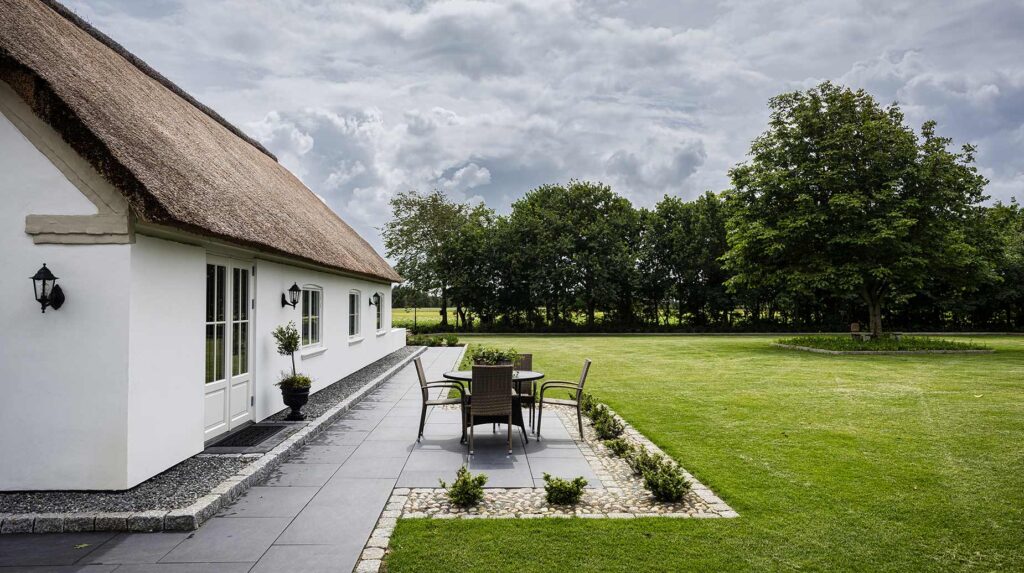 Anlagt terrasse med flisebelægning og bede op til stråtækt hus, med velholdt græsplæne og beplantning i baggrunden.