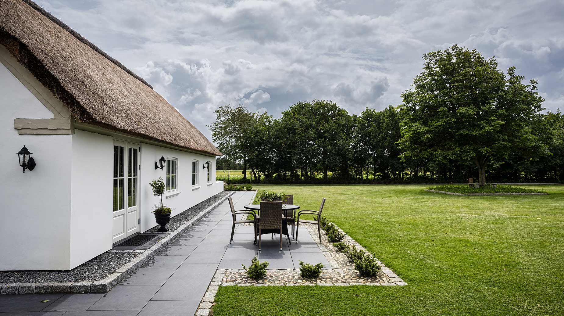 Anlagt terrasse med flisebelægning og bede op til stråtækt hus, med velholdt græsplæne og beplantning i baggrunden.