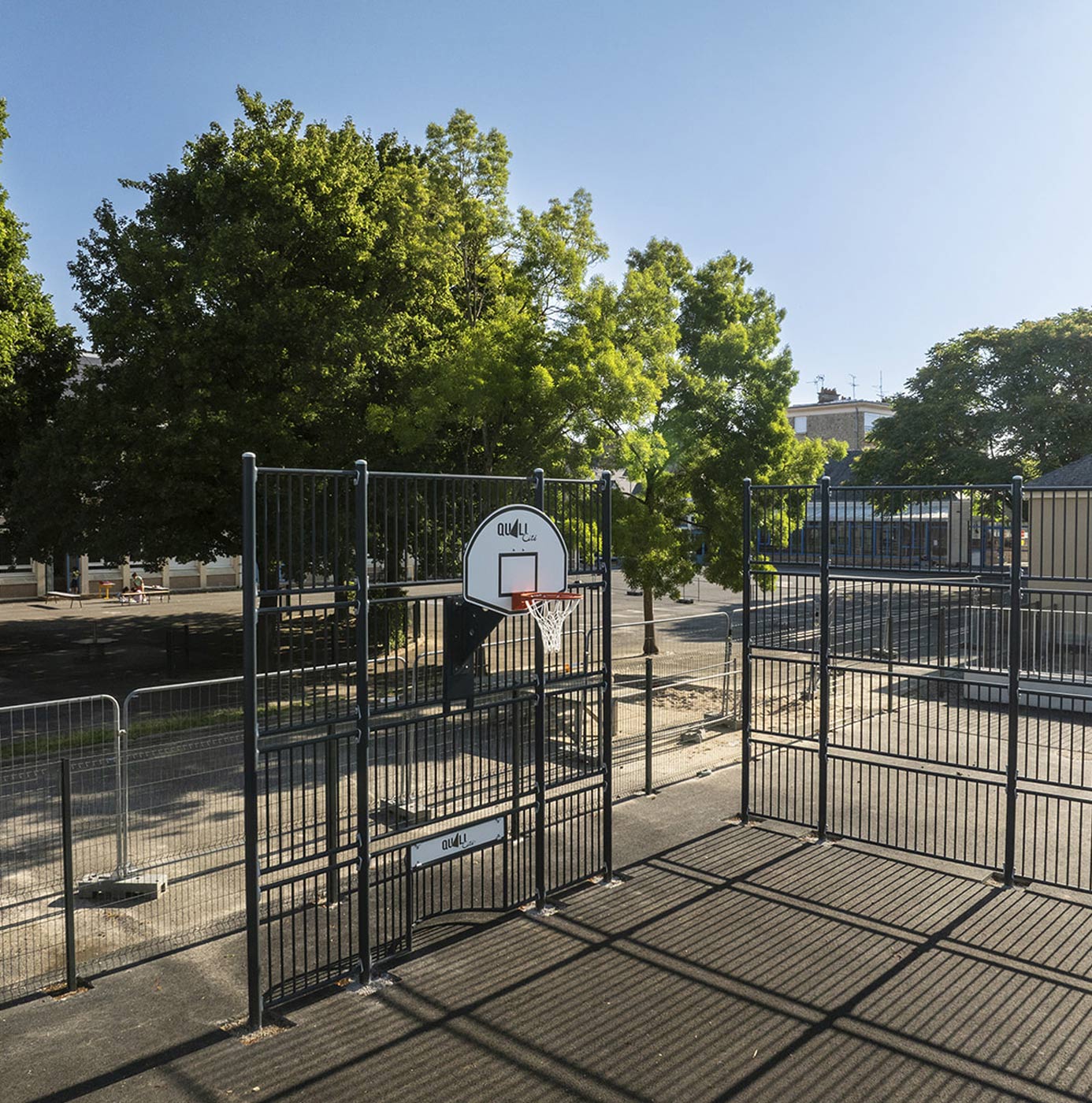 Indhegnet multibane med basketkurv omgivet af nyplantede træer og grønne kroner ved skolegård.