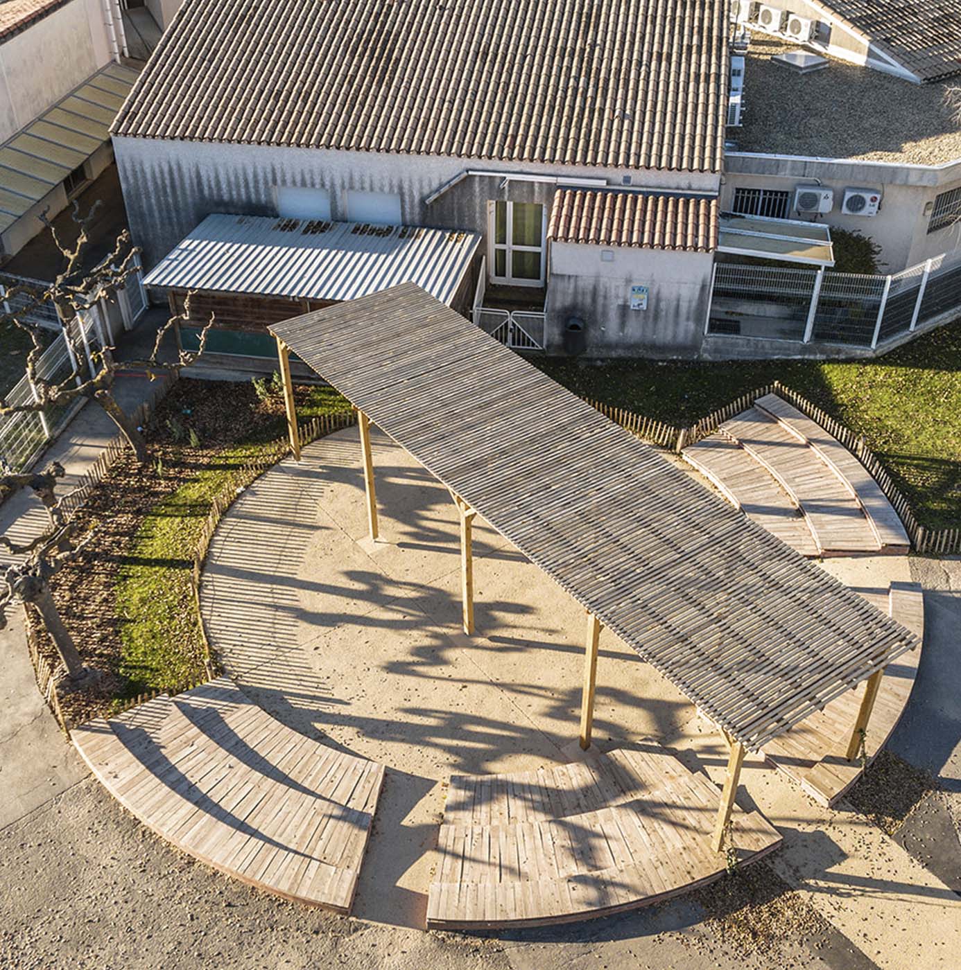 Luftfoto af cirkulær udendørs opholdsplads med træpergola og amfiteaterlignende trædæk ved skolegård.