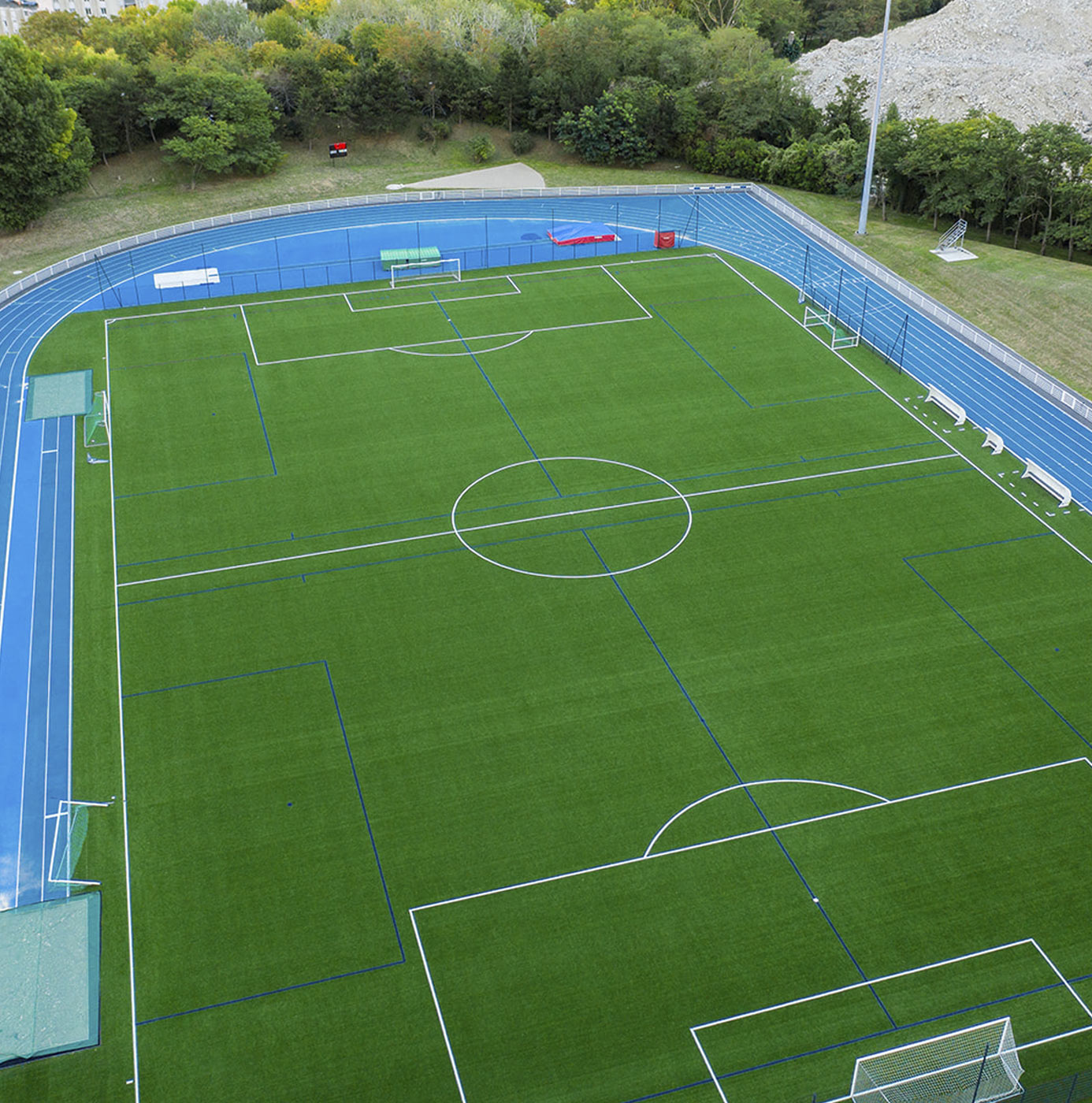 Luftfoto af kunstgræsbane med atletikbane omkring, anlagt ved sportsfacilitet omgivet af træer.
