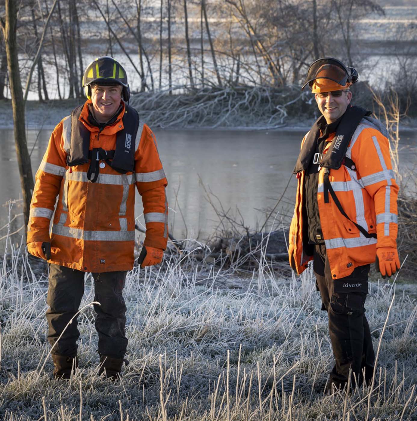 To idverde-medarbejdere i sikkerhedsudstyr og redningsveste ved en frostklædt å.