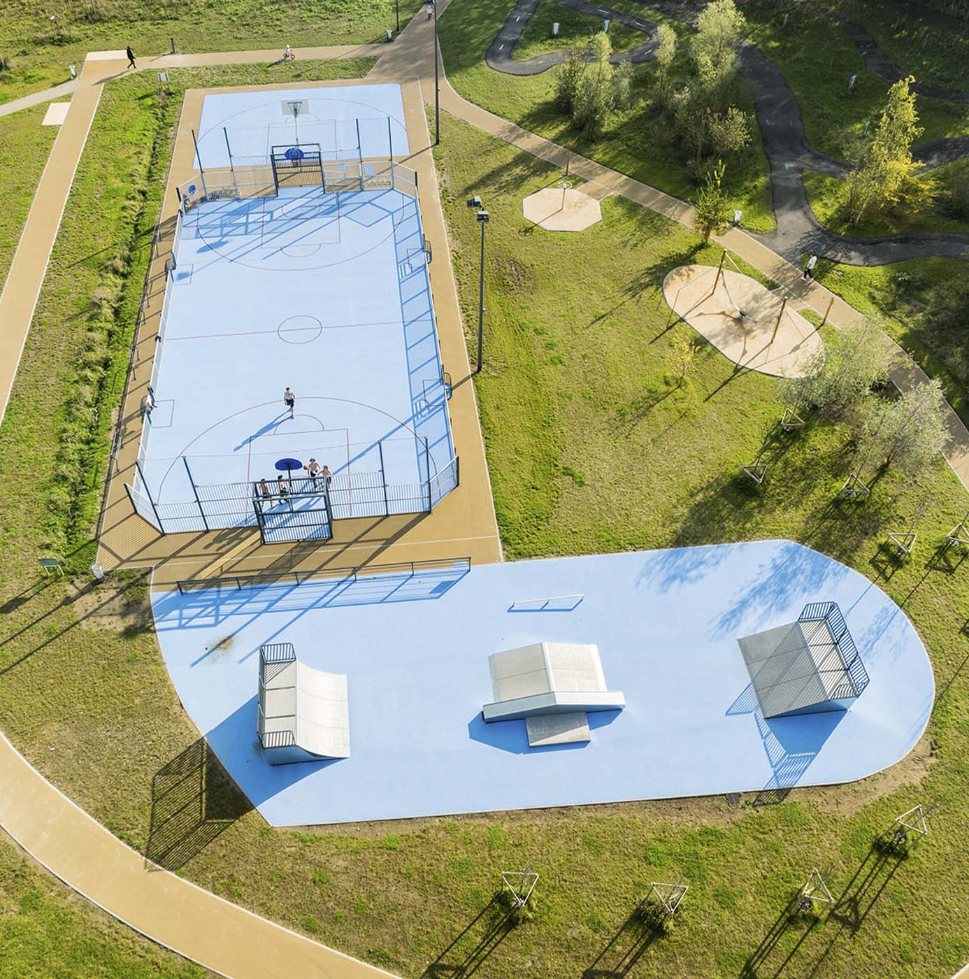 Luftfoto af anlagt idrætsområde med basketbane, skaterbane og legeplads omgivet af grønne arealer og stiforbindelser.