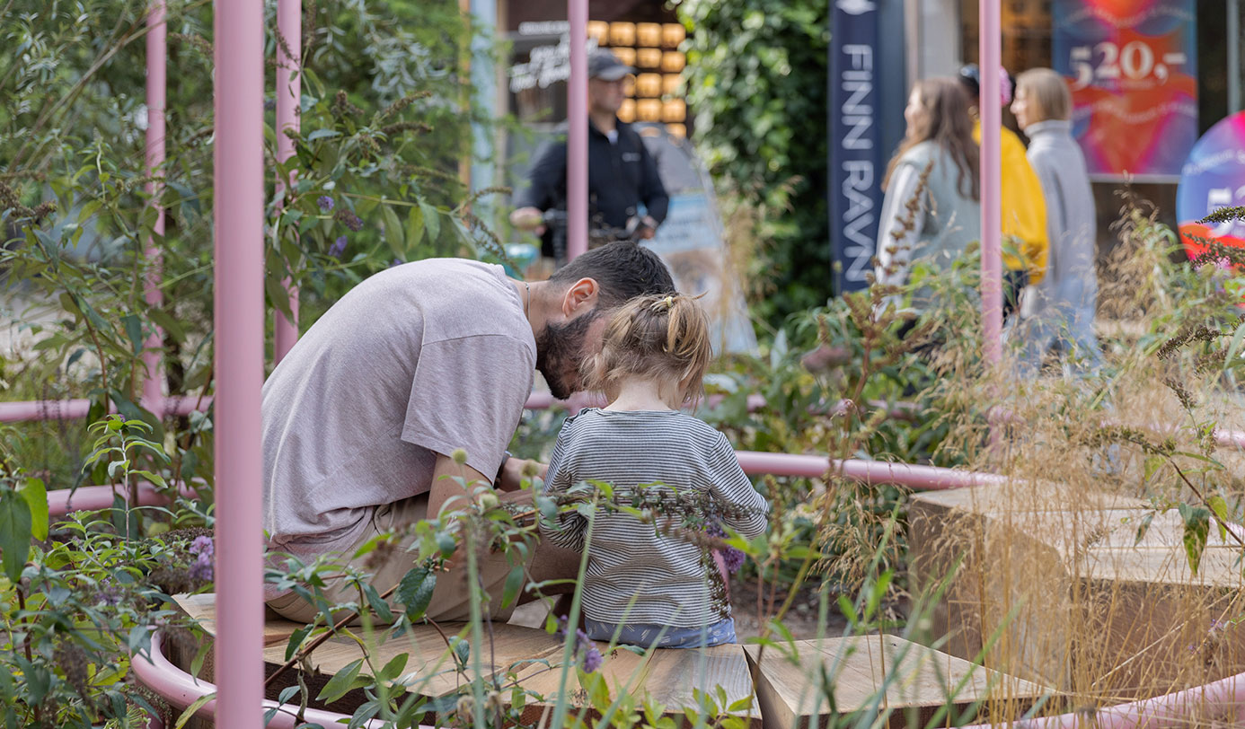 Voksen og barn kigger på planter i bydelsbeplantning med pink stålkonstruktion i bymiljø.