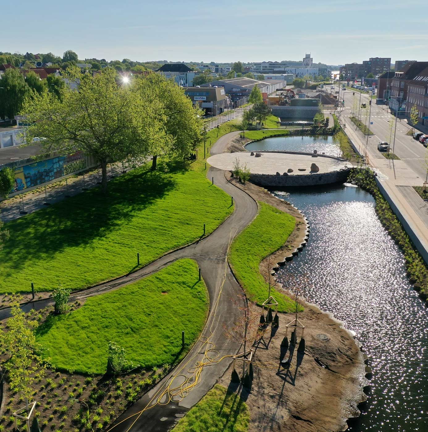 Nyanlagt parkområde langs vandløb med slyngede stier, græsplæner og nyplantede træer i byområde.